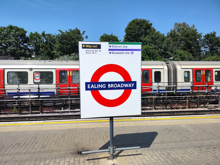 A detailed view of a train station platform in daylight with a large white sign displaying the Ealing Broadway underground station name in bold blue and red letters, with directional arrows pointing t