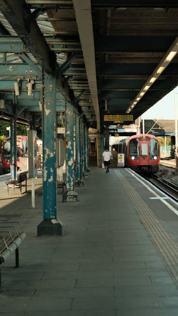 A detailed view of a train station platform in daylight with a large white sign displaying the Ealing Broadway underground station name in bold blue and red letters, with directional arrows pointing t