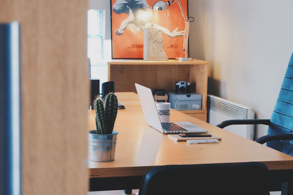 View of a workspace through a partially open door, showing a wooden desk with a silver potted cactus plant in the foreground, alongside an open laptop, a notepad, and a pen. Behind the desk, there is a wooden shelving unit with a video camera, a printer, and a small stack of paper. A framed poster of a person in motion is mounted on the wall above the shelf, and a desk lamp is positioned on the top of the shelf. To the right, a blue office chair with armrests is visible, and a white radiator is installed on the wall. The room features natural light coming from a window on the left side, illuminating the workspace, which appears organized and ready for professional tasks. This setting depicts an office environment prepared for a furniture transport or packing and moving activities, representative of the services offered by Colliers Wood Removals in facilitating house and office relocations.