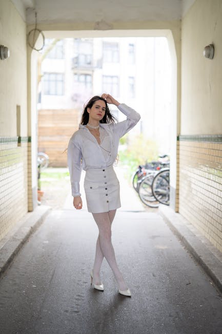 A young woman with long dark hair stands in a covered outdoor passageway, with sunlight illuminating the area behind her. She is wearing a white button-up shirt and a white skirt, with light-colored tights and white high heels. Her left arm is raised to her head as she gently touches her hair, and she has a neutral, relaxed expression. The passageway has beige brick walls with a green and brown tiled trim along the lower half, and two round wall-mounted lights are visible on either side. In the background, there is an open space with a wooden fence, some bicycles parked against it, and a hint of greenery. This scene presents a casual, urban setting, and while no moving or relocation activities are depicted here, the image aligns with themes of personal style and daily life in a residential area.