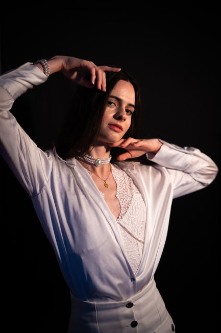 A young woman with shoulder-length dark hair and light skin tone, posed against a black background, is wearing a white satin blouse with lace detailing on the front and a collar. She is accessorized with layered necklaces, including a pearl choker and a small gold pendant, as well as a bracelet on her right wrist. She has a confident expression, with her left hand resting near her neck and her right hand raised above her head, touching her hair. The lighting highlights her face and upper body, creating a contrast with the dark backdrop. The overall styling and pose suggest a fashion or artistic portrait setting.